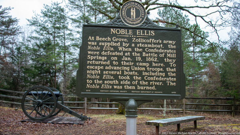 A Noble Ellis marker stands beside a field cannon and bench among trees at the battlefield.