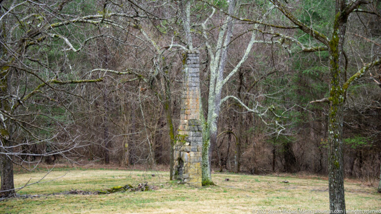 A narrow stacked-stone pillar rises among leafless trees beside a grassy clearing.