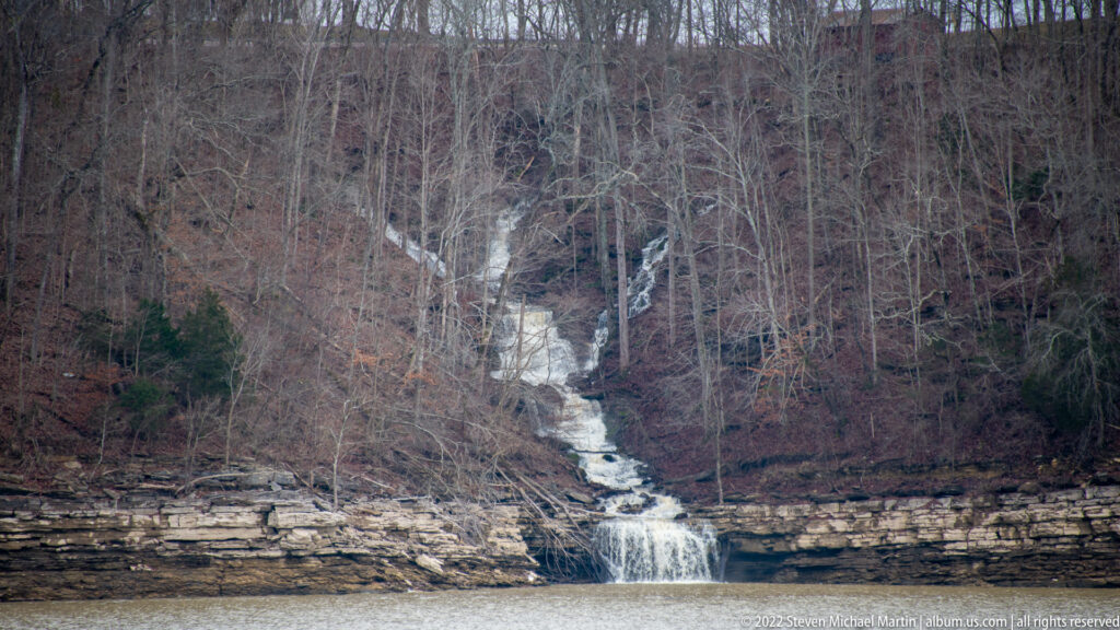 A narrow waterfall descends over pale rock ledges through a leafless wooded ravine into a pool.