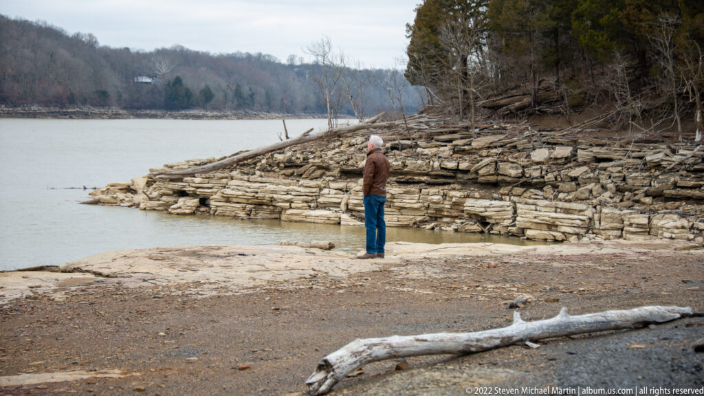 A visitor stands on a limestone shelf beside the Cumberland River, facing layered rock and wooded banks.