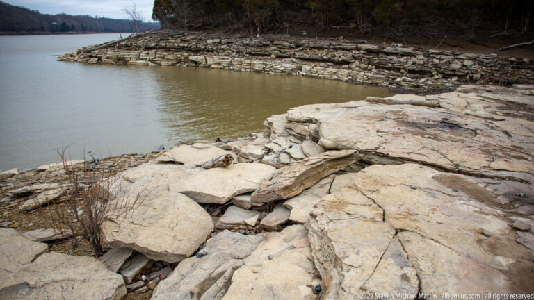 Layered limestone shelves curve around still river water beneath a wooded bank.