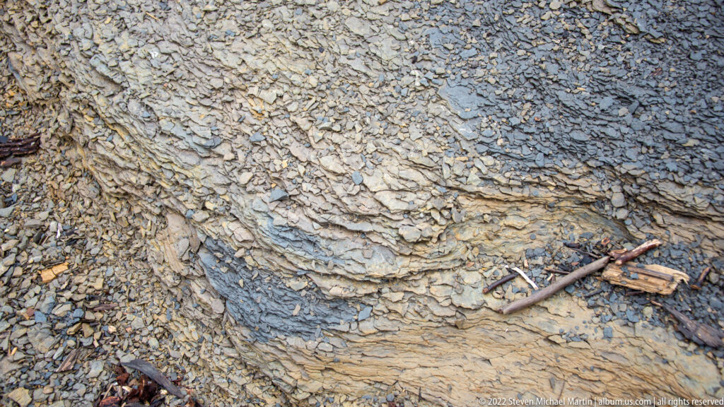A close view of folded, layered rock shows a branch across the exposed bank beside a small patch of water.