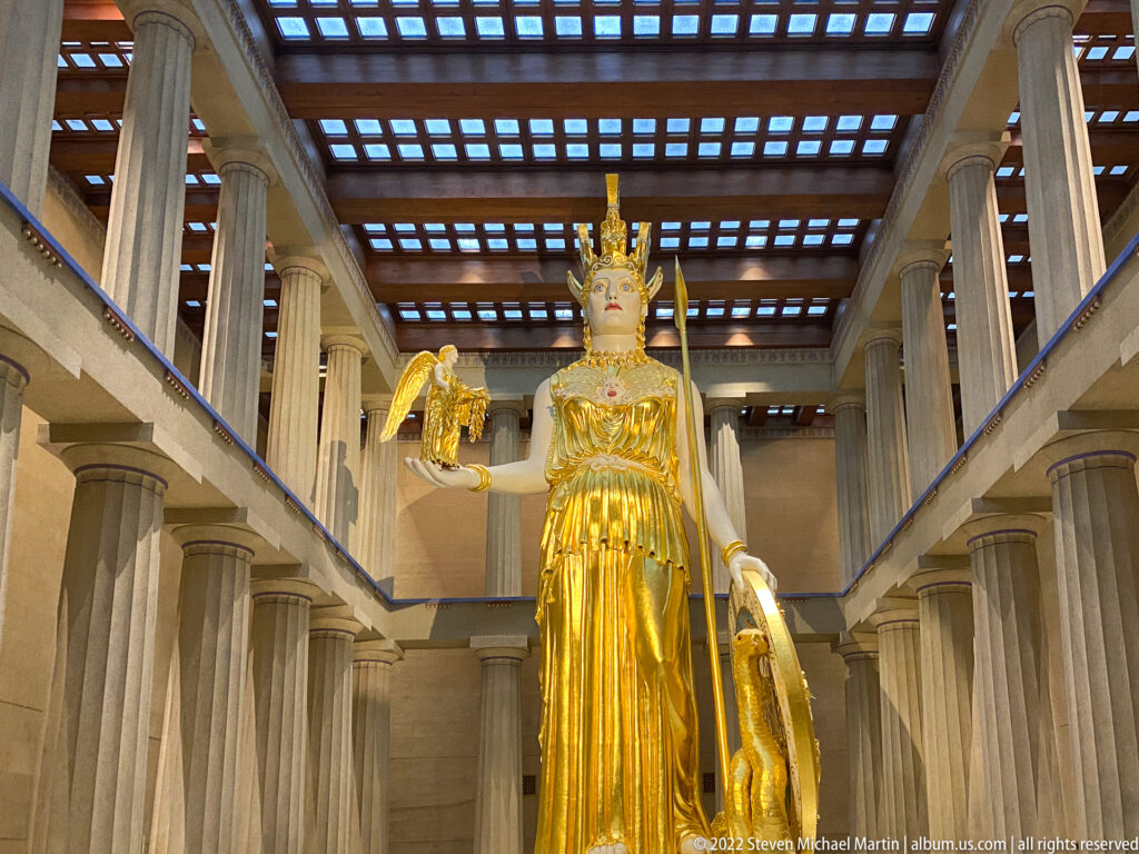 A gilded Athena statue stands between columns beneath a coffered ceiling, holding a small figure and shield.