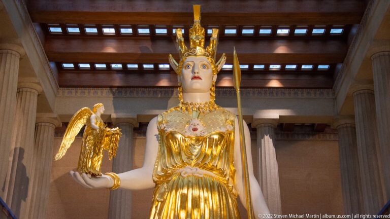Athena’s gilded upper body and helmet fill the frame as she holds a winged figure.