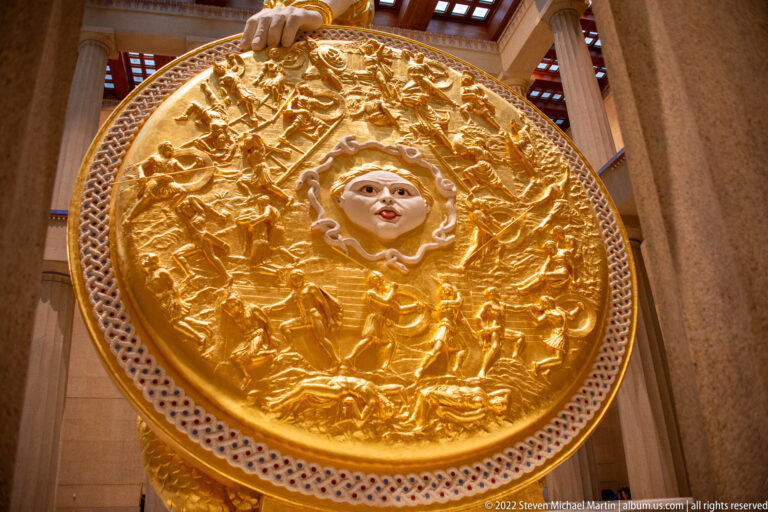 A central face and many raised figures cover Athena’s circular gold shield, edged with a beaded border.