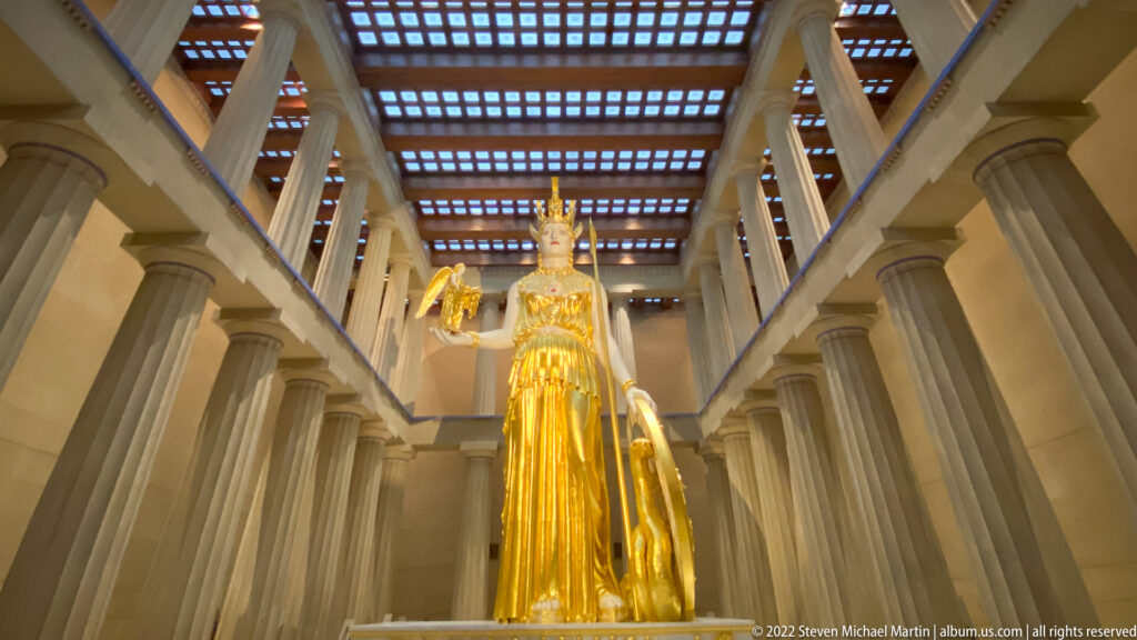 Athena stands at the center of a columned hall beneath a patterned timber ceiling.