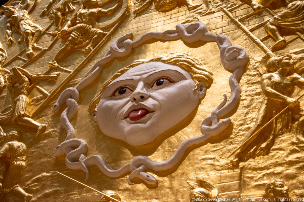A sculpted face with red lips sits inside a curling raised border on a gold shield.