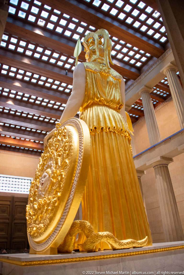The back of Athena’s gilded robe and helmet is visible beside the edge of her shield.