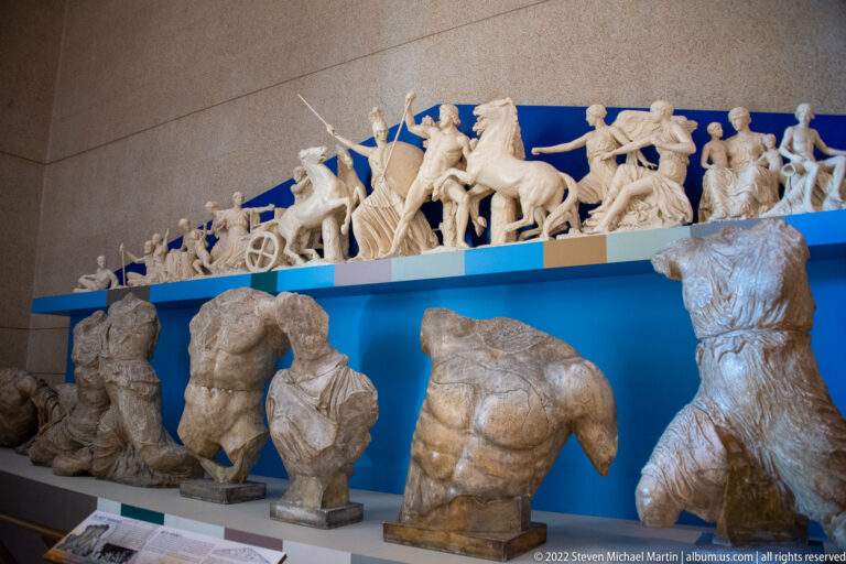 Headless stone torsos stand on individual plinths below a long sculpted frieze against blue.