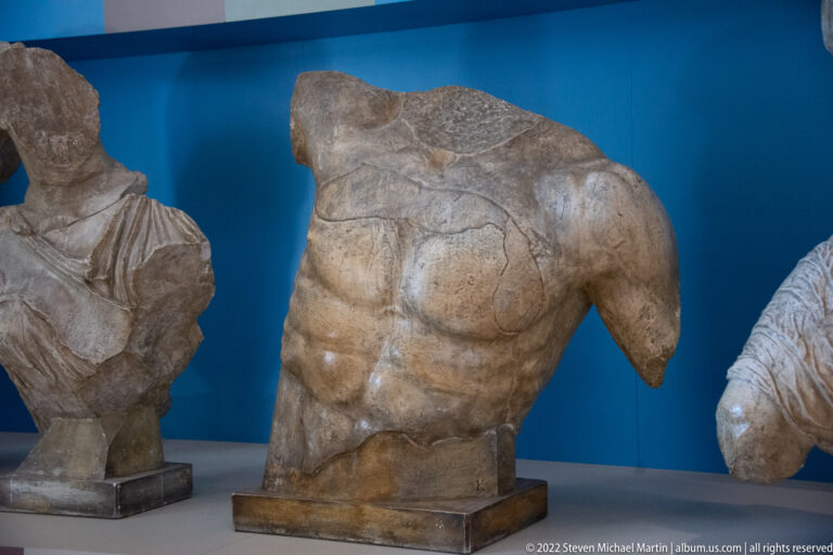 A headless stone torso with defined abdominal muscles stands on a dark base.