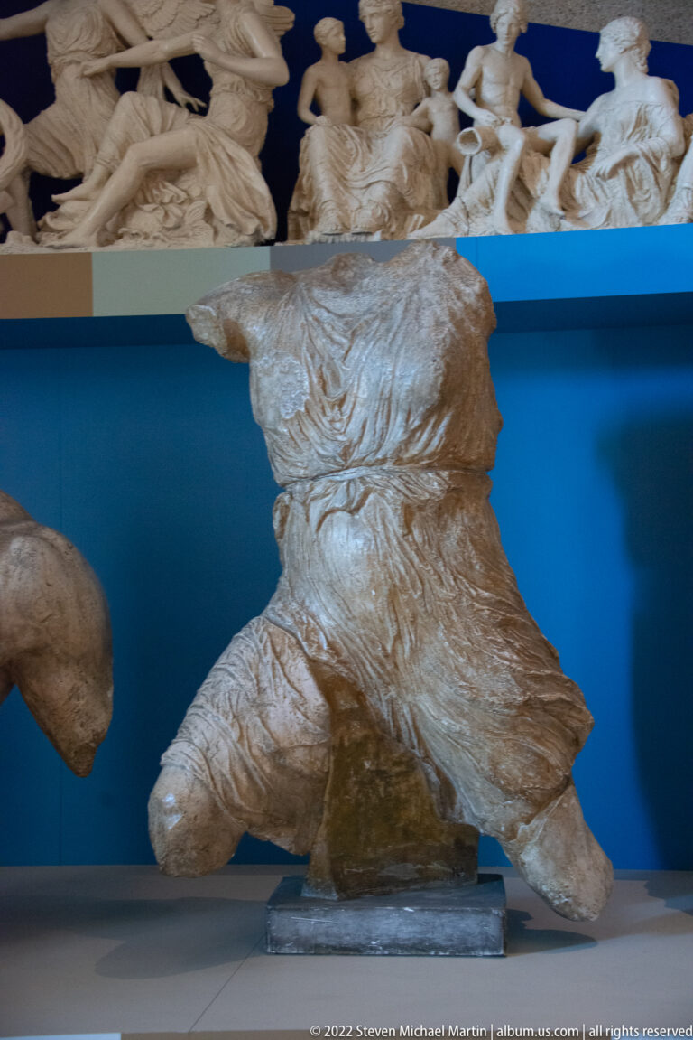 A pale draped sculpture fragment with bent legs stands below a row of small figures.