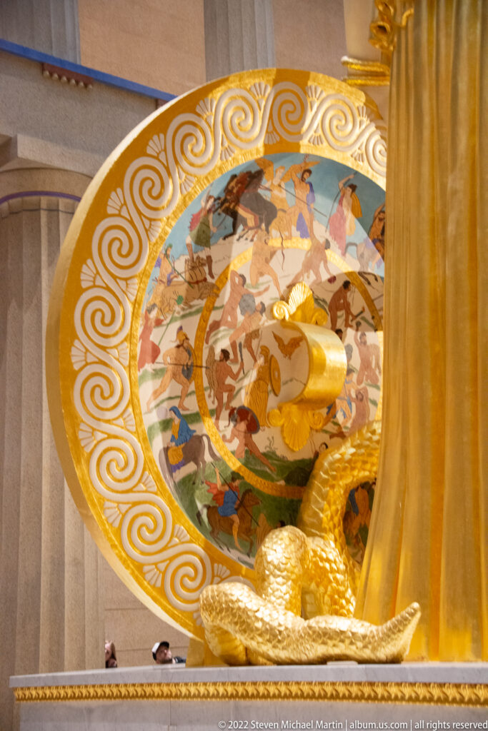 A circular painted scene with human figures is set within a gold ornamental shield.
