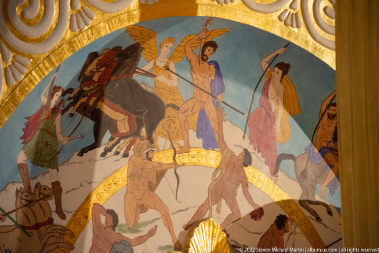 Painted human figures and a chariot appear inside a curved gold border on the shield.