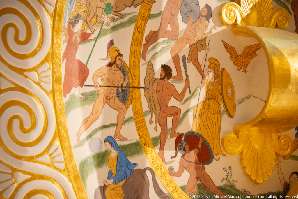 Painted figures with spears and shields face one another inside a gold-bordered scene.