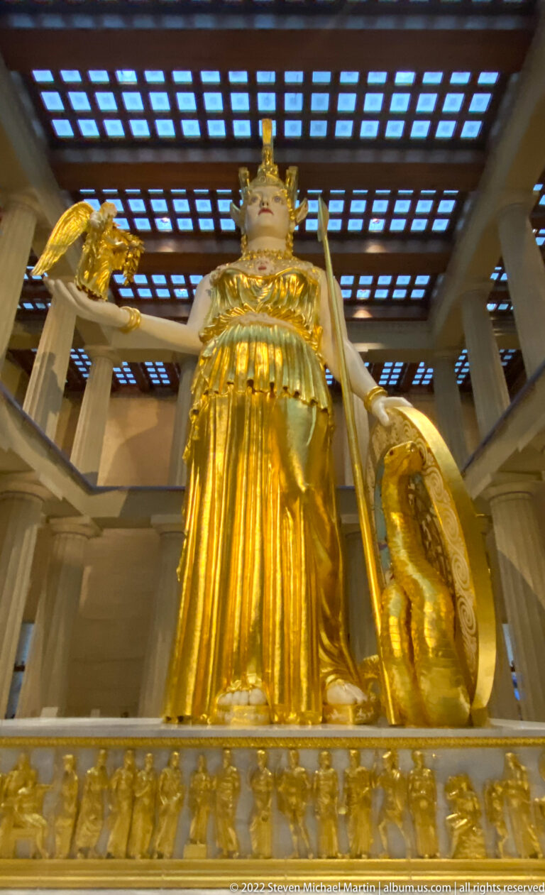 A low view of Athena shows her gilded armor, spear, round shield, and sculpted base.