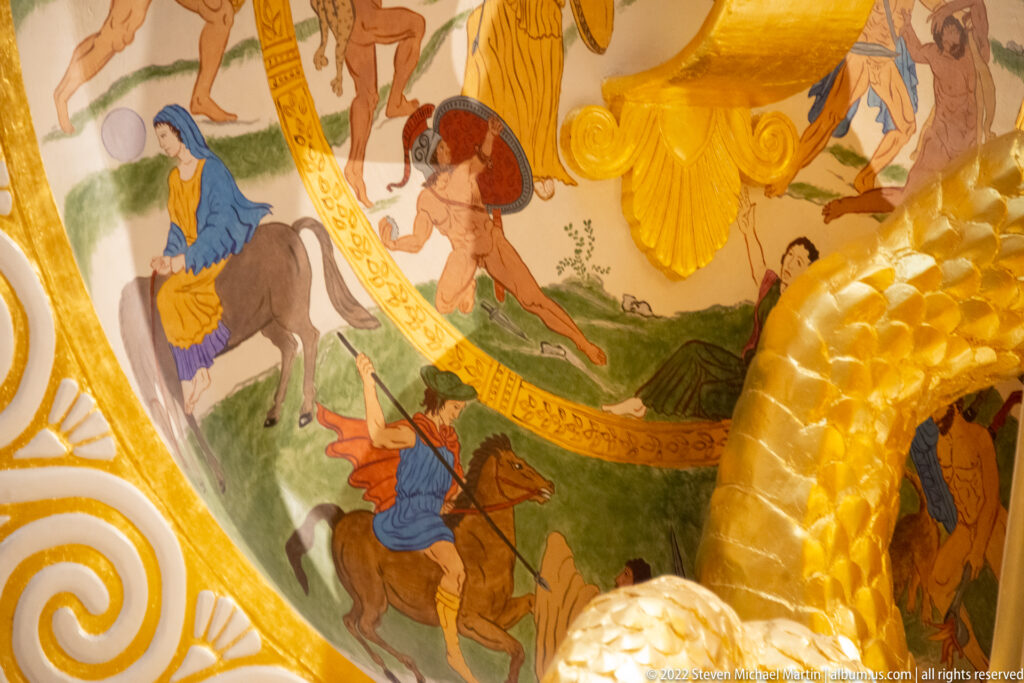 A painted rider on horseback appears beside other figures within the shield’s curved gold frame.