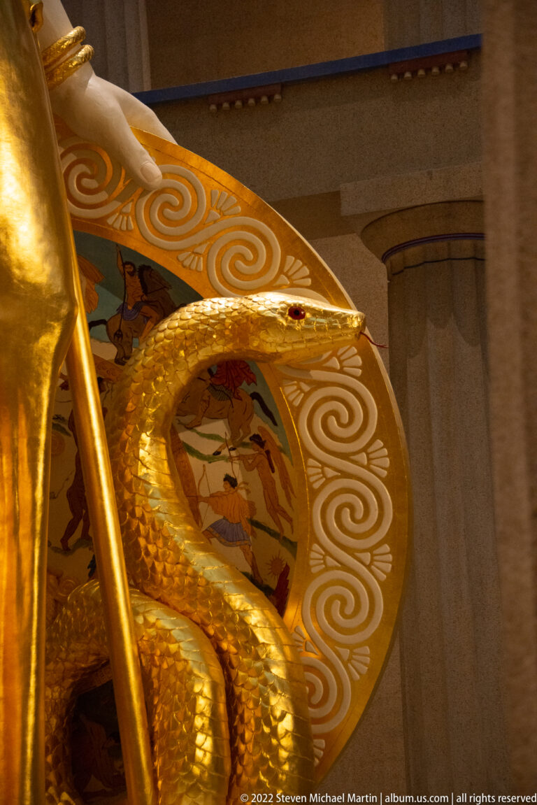 Gold serpent coils across a decorated shield with spiral motifs and a small painted scene behind it.