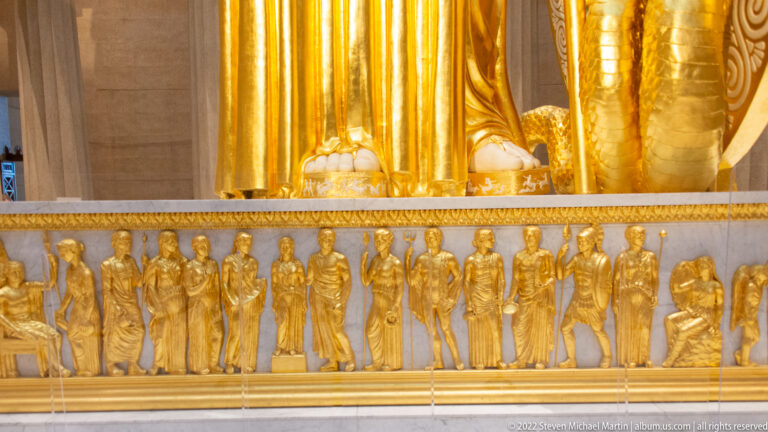 Row of small gilded figures in varied poses runs along the lower edge of a monumental gold statue.