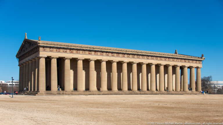 A long colonnaded temple stands across an open lawn beneath a clear blue sky.