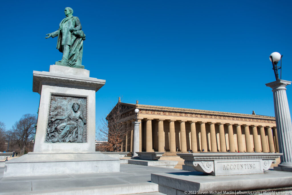 A standing bronze-colored statue on a relief-decorated pedestal stands before the Parthenon.