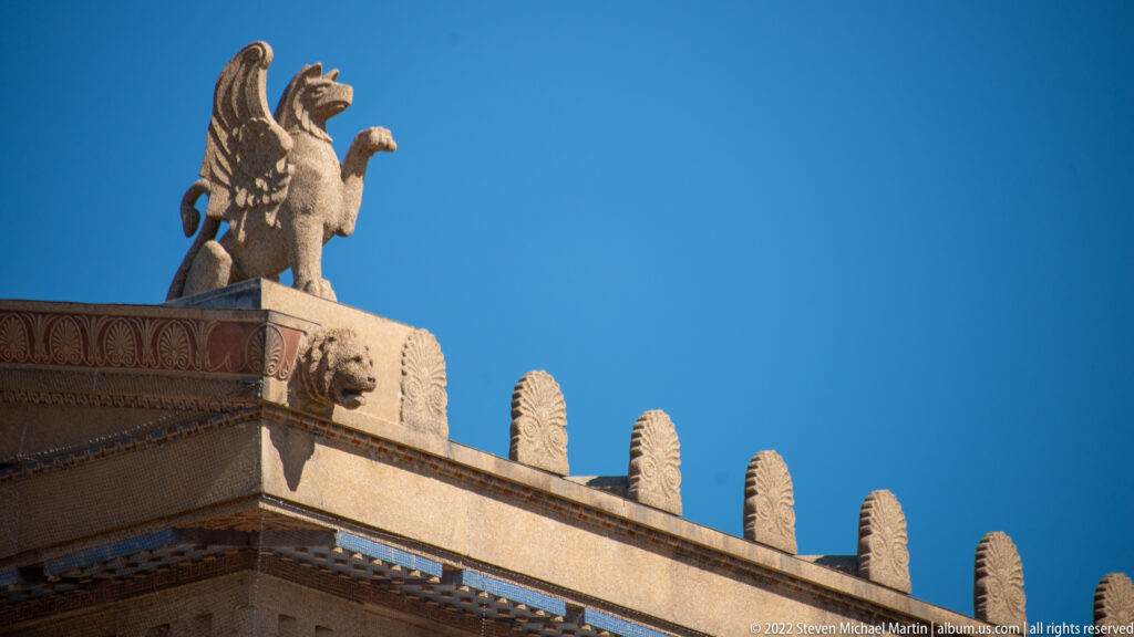 A stone winged creature sits at the temple roof corner above a carved frieze.