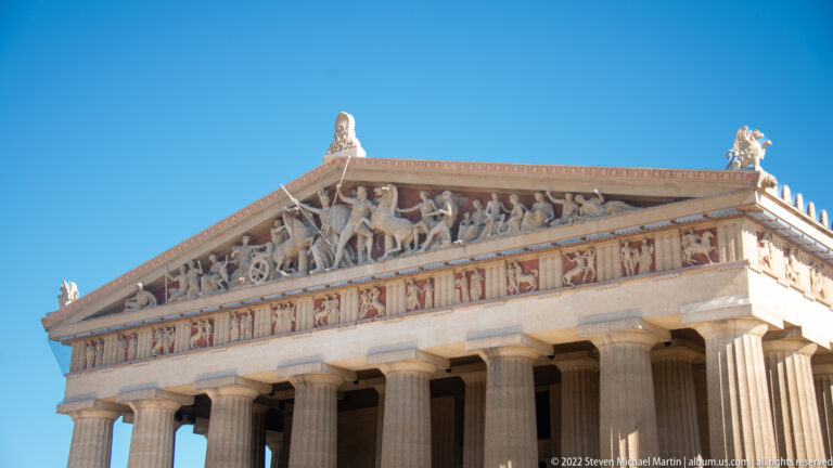Pale sculpted figures fill a triangular pediment above a painted frieze and columns.