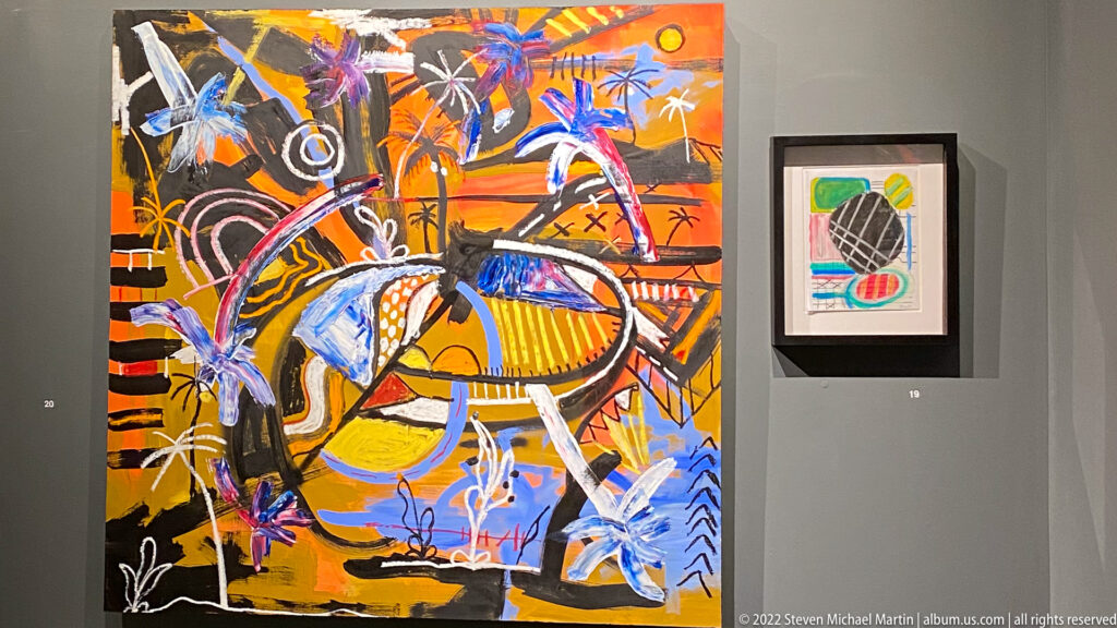 Large abstract painting with bright orange, yellow, blue, and black marks hangs beside a smaller framed artwork.