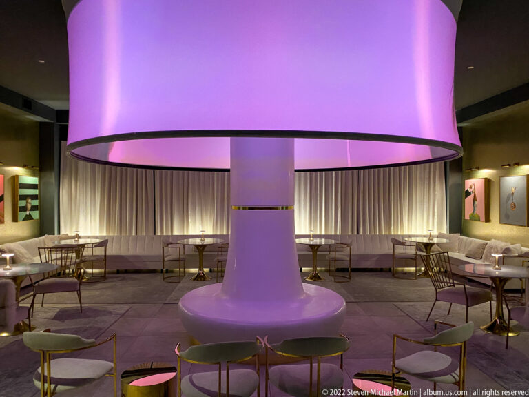 A glowing pink circular canopy and central column hang above chairs and tables in a curtained lounge.