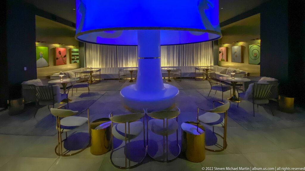 Blue light illuminates a circular canopy and central column above a ring of chairs and tables.