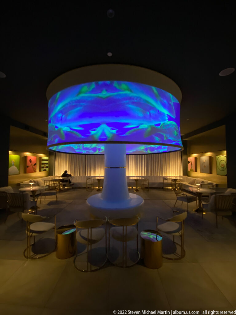 A luminous circular canopy displays blue, green, and violet patterns above lounge seating.