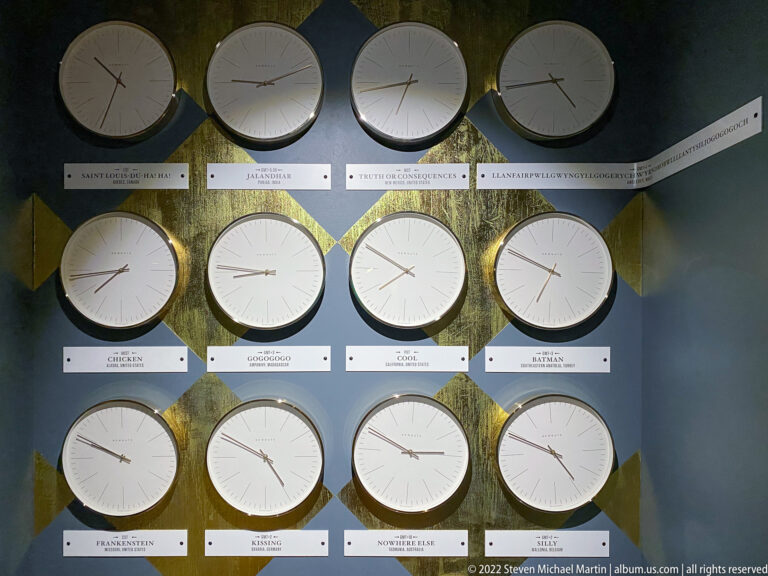 Twelve pale round clocks with caption strips arranged in three rows on a dark wall.