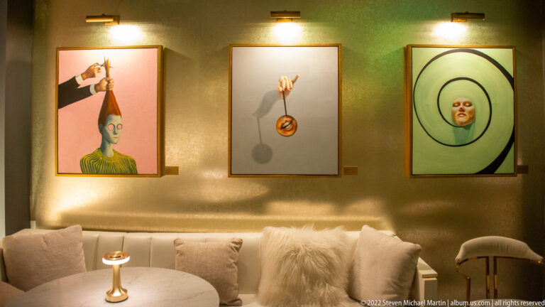 Three surreal paintings hang above a pale sofa, including stylized faces and a suspended circular form.