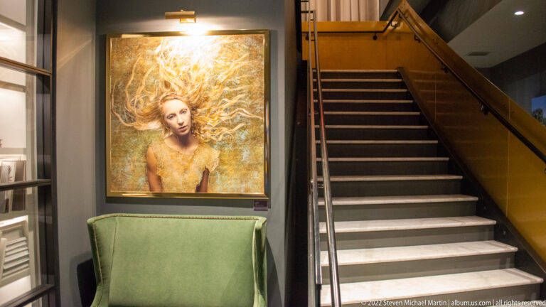 A gold-toned portrait of a woman hangs above a green chair beside a staircase.