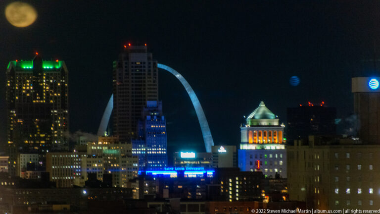 The Gateway Arch rises behind illuminated downtown St. Louis buildings against a dark night sky.