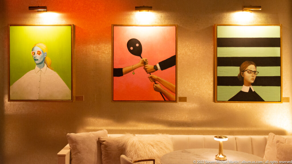 Three colorful paintings of figures and faces hang above a pale sofa and small round table.