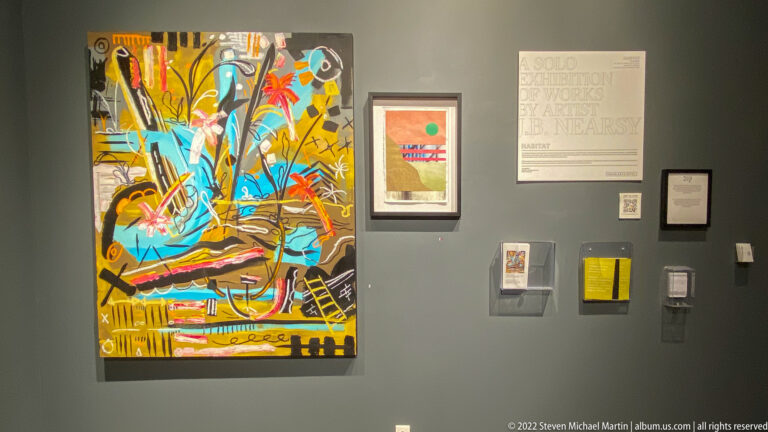 A large colorful abstract canvas hangs beside framed works, small display cases, and interpretive text on a gray wall.