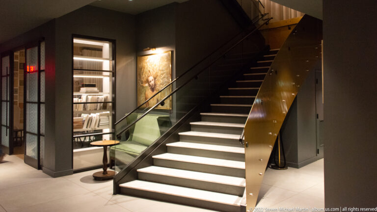 A staircase rises beside a bookcase, green chair, small table, and framed portrait in a dark interior.