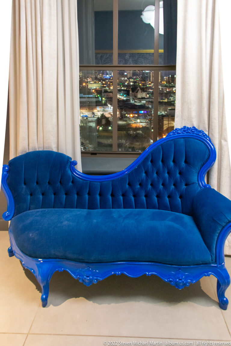 A blue tufted velvet sofa stands before a curtained window overlooking city lights at night.
