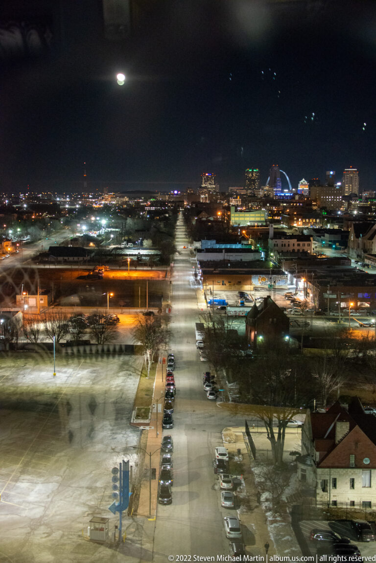 An elevated nighttime view shows parked cars, a lit access lane, low buildings, and distant downtown lights.