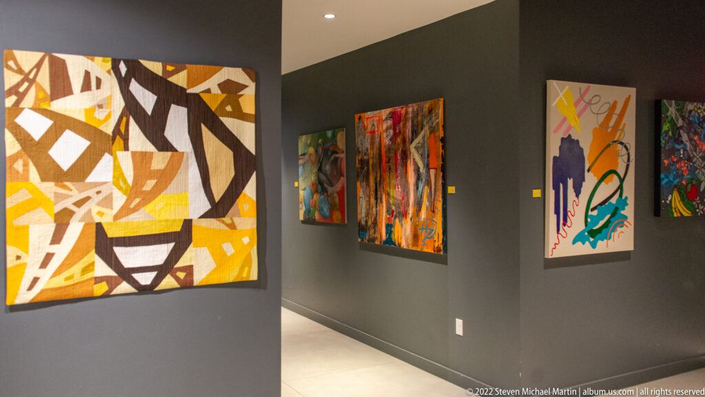 A large geometric painting and several colorful works line adjoining gray gallery walls.