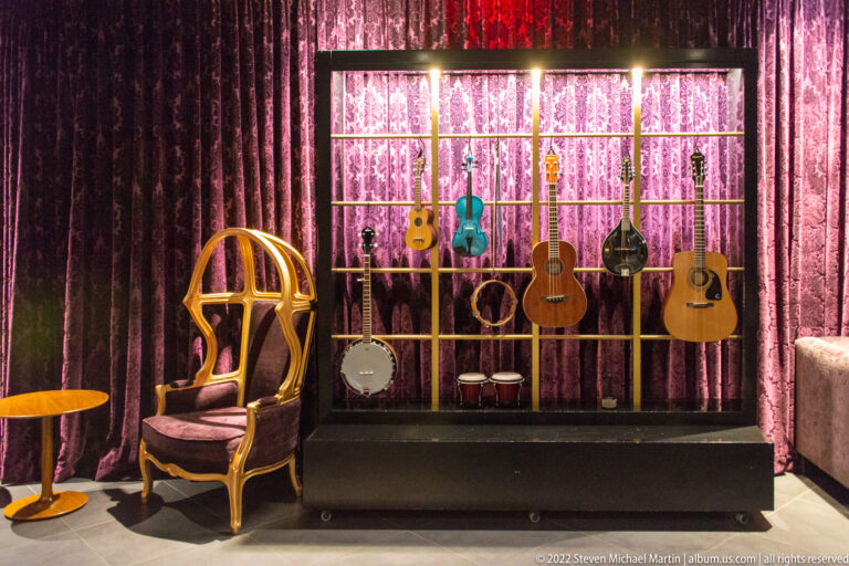 Guitars and a banjo hang against a pink-lit curtain behind a gold-colored chair and small table.