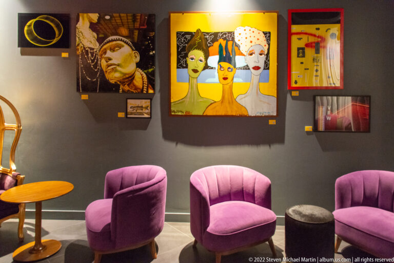 Pink upholstered chairs face a wall of artworks, including a large painting of three stylized faces.