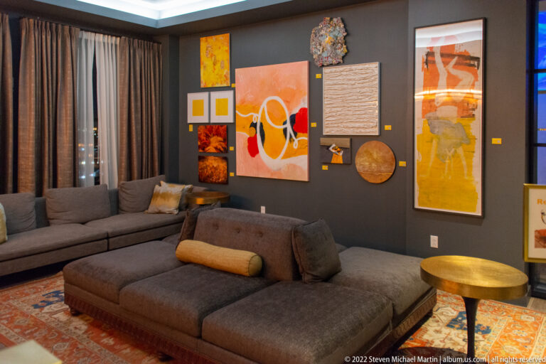 A gray sectional and brown chaise face a wall of colorful paintings and objects above a patterned rug.