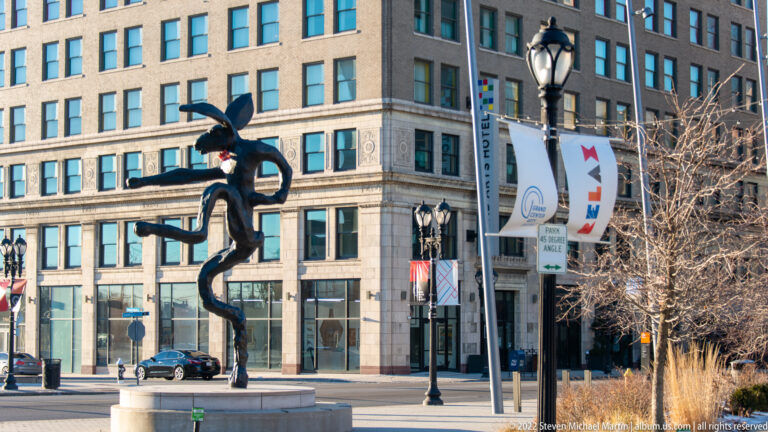 A dark twisting figure-like sculpture stands beside a street lamp and banners in front of a downtown building.