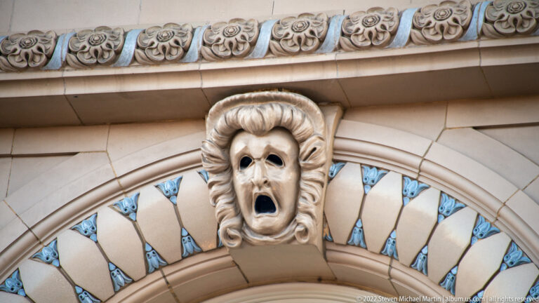 An open-mouthed carved face projects from a pale stone facade surrounded by ornamental details.