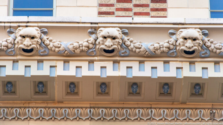 Three sculpted faces and garland forms repeat across a facade above a row of recessed decorative panels.