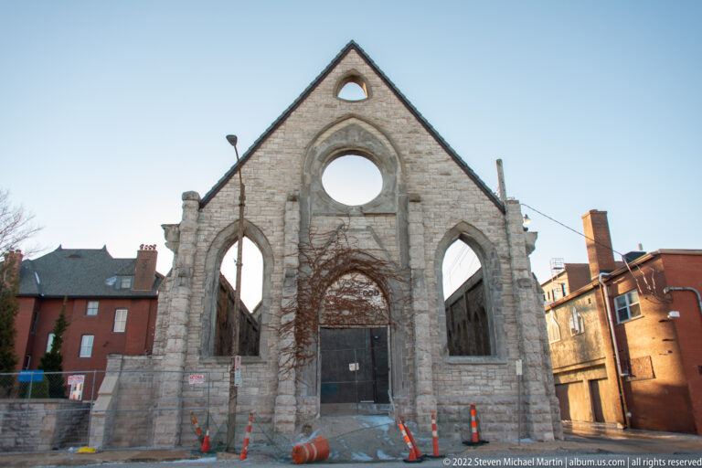 Weathered stone Gothic façade with empty circular and pointed openings between brick buildings.