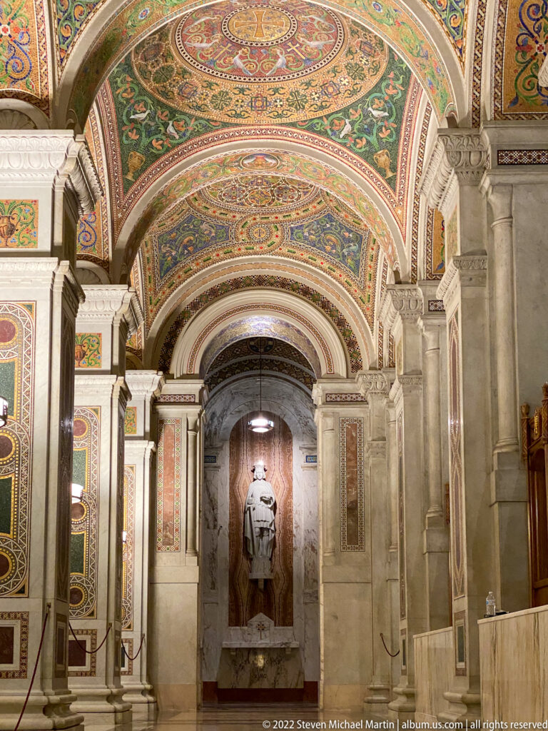 Ornate church nave with mosaic-covered vaults, stone piers and a statue in a distant niche.