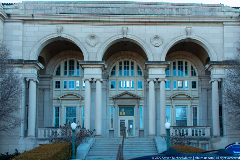 Symmetrical pale-stone façade with three arched entrances, columns, stairs and upper windows.