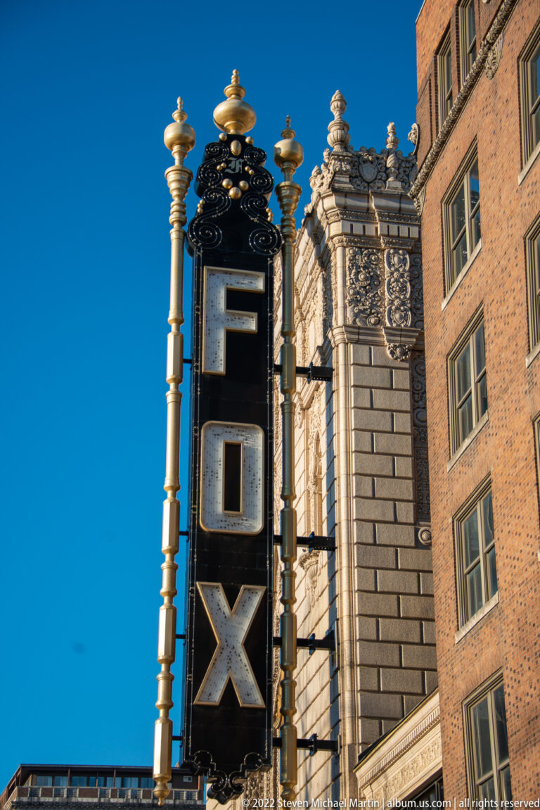 Stacked “FOX” sign rises beside an ornate stone theatre façade under a clear blue sky.